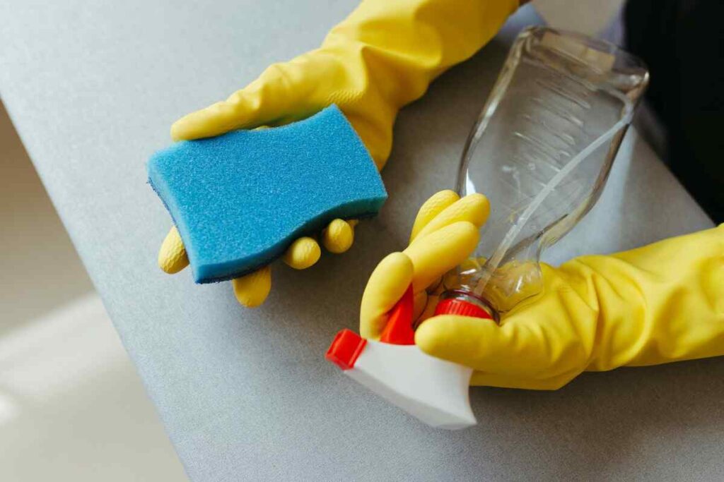 Handen met gele schoonmaakhandschoenen houden een blauwe spons en een sprayfles vast voor het schoonmaken van een airfryer.